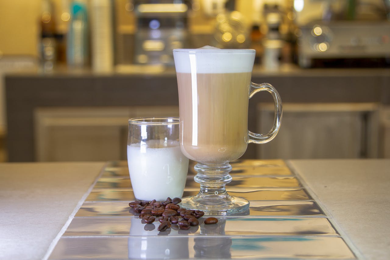 A stylish latte served in a glass mug with a glass of milk and coffee beans on the table in a cozy cafe setting.