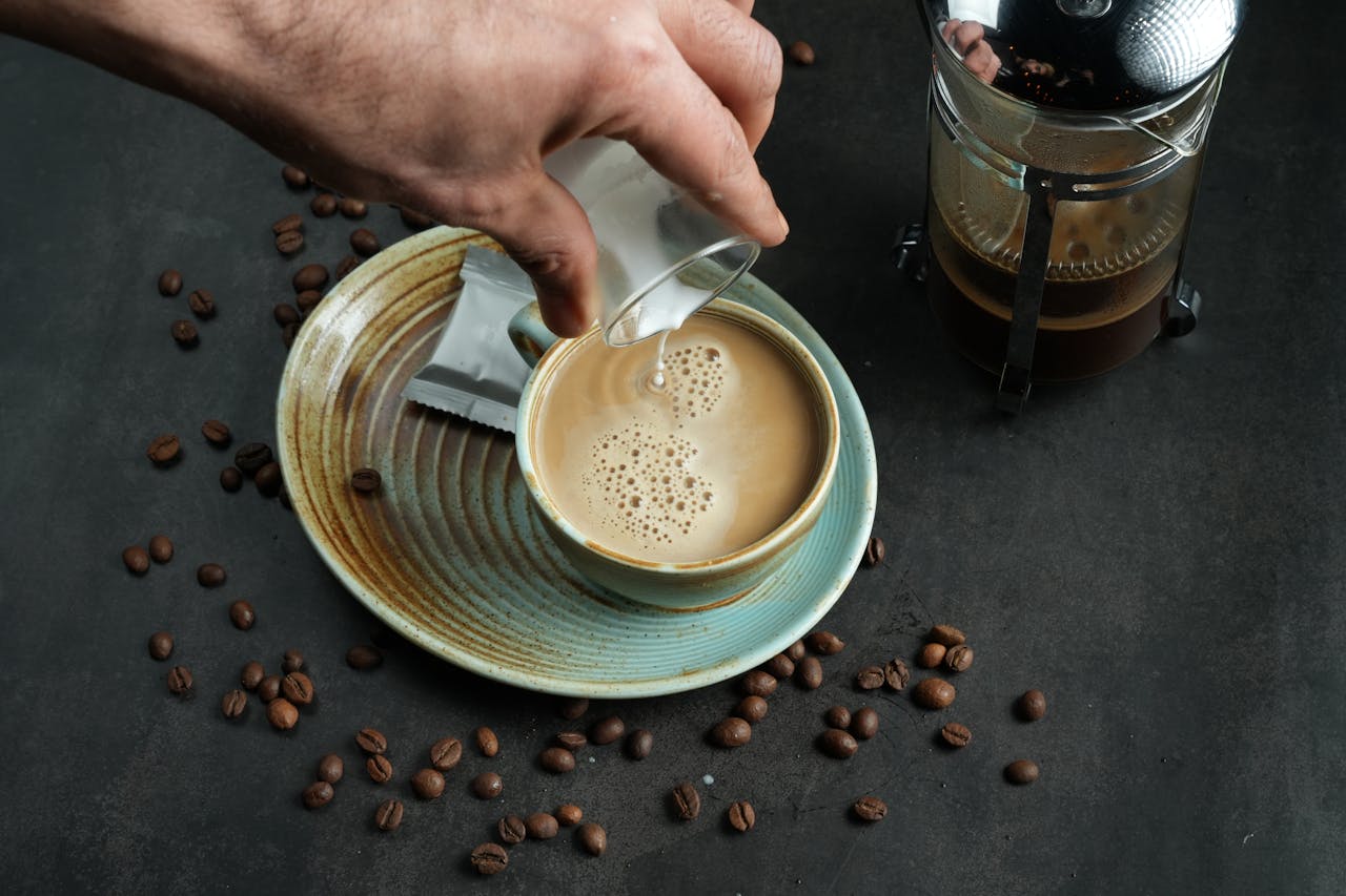 Close-up of coffee with milk pouring and French press on a dark surface.